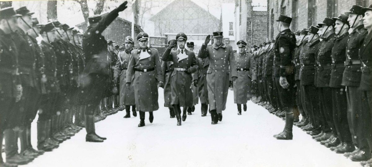 Svart-kvitt-bilete. Ei gruppe menn i tyske uniformar går opp ei gate. Langs vegkantanen på begge isder står Rikshirden i  akt. Ein mann har armen heva i Hitler-helsing.