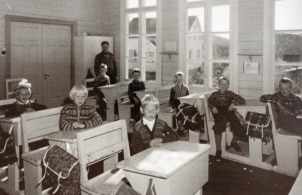 Lærer og 8 skolebarn sitter ved skolepultene sine i klasserommet og ser mot fotografen