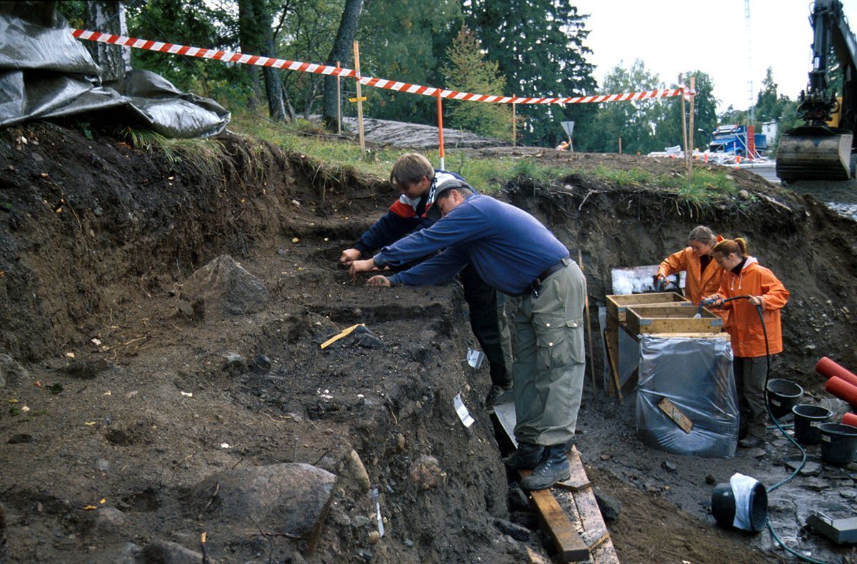 Fotografi fra utgraving: to menn i forgrunn graver i jordparti, mens to kvinner i bakgrunn står ved spylekar