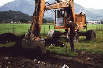 Her er arkeologen Trond Løken i gang med å fjerne åkerjord ved hjelp av gravemaskin på Forsandmoen i Rogaland, for å avdekke strukturer i undergrunnen. Denne metoden revolusjonerte bosetningsarkeologien . Foto: Asbjørn Simonsen/Arkeologisk museum, Universitetet i Stavanger/ CC BY-NC-ND 3.0