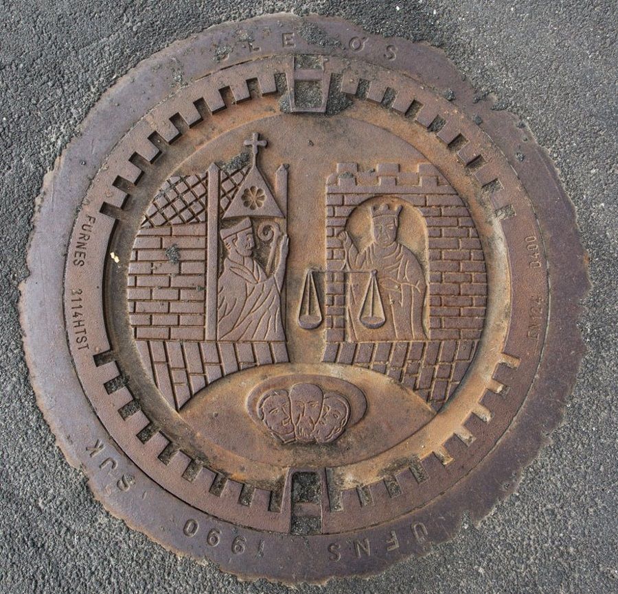 Fargefoto av eit kumlokk me asfaltkant rundt. Motivet for kumlokket er to skikkelsar som sit i kvart sitt borgtårn.