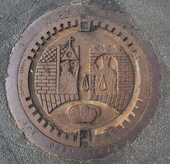 I Trondheims byvåpen fra 1200-tallet kan se en konge i et tårn i en borg på den ene siden, og en erkebiskop i et tårn i en katedral på den andre. De to skikkelsene står på samme linje , den ene er altså ikke hevet over den andre, noe som symboliserer todelingen av statsmakten. Her ser vi det gamle byvåpenet gjengitt i et kumlokk fra samlingen til Gosen skole i Stavanger. Foto: Knut Roberto Ås/ Kumlokk kulturprosjekt – Gosen skole