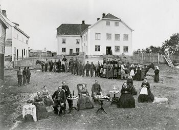 Gårdbrukeren på storgården Nedre Rogneby på Østre Toten, med familie, oppstilt i forgrunnen. Huslæreren sitter med familien, mens husmenn, dagarbeidere, tjenestefolk og legdslemmer (fattige som ble forsørget ved å vandre fra gård til gård). Fotografiet er tatt i 1869 og demonstrerer en vanlig fotografisk praksis på 1800-tallet: Innendørs fotografi var vanskelig, og derfor ble eiendeler som vitnet om sosial status, gjerne stilt opp utendørs. Les mer om dagliglivet og om hvordan levekår ble forbedret på 1800-tallet . Foto: Per Adolf Thoren/Mjøsmuseet