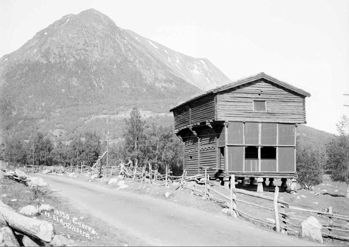 Svart/hvitt-foto av stort stabbur langs landevei, fjell i bakgrunn