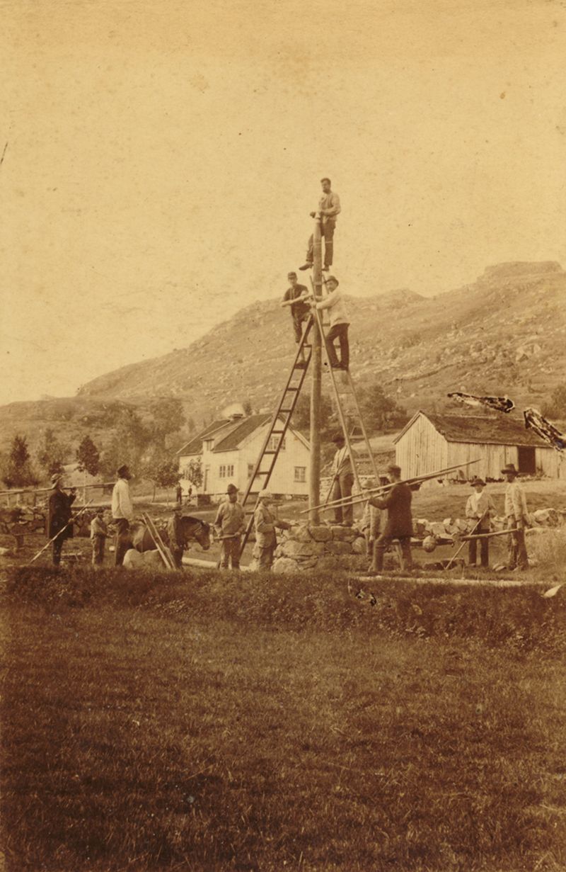 Fotografi som viser byggingen av en telegraflinje: to menn på stiger opp langs en mast, i landlige omgivelser