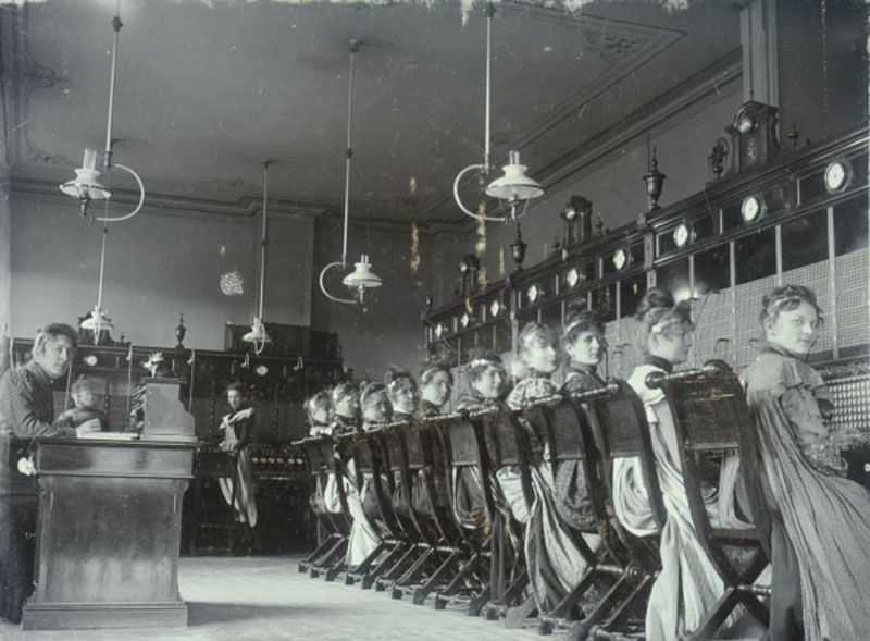 Fotografi av kvinner som sitter på rad langs telegrafapparater, med ryggen mot fotografen, men tittende over skuldra.