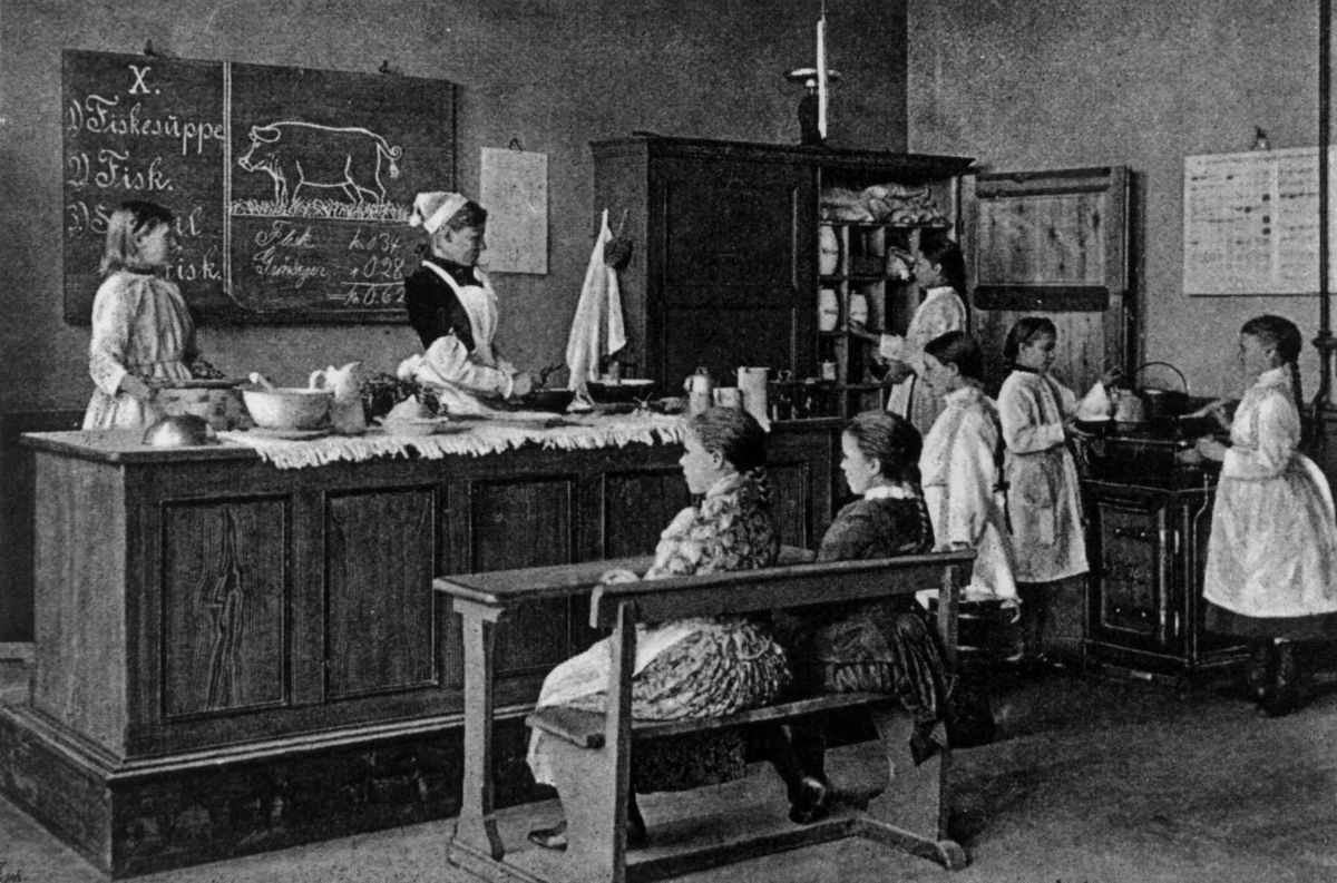 Foto som viser skolekjøkkenundervisning: jenter blir undervist av kvinne ved kateter