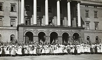 Skikken med å hilse barnetoget fra slottsbalkongen på 17. mai ble innstiftet av kong Haakon og dronning Maud. Her ser vi det første barne­toget som ble hilset av den nye kongefamilien på slottsbalkongen i 1906. Foto: ukjent/Aktieselskapet Norsk Kunst /Nasjonalbiblioteket/ CC BY SA