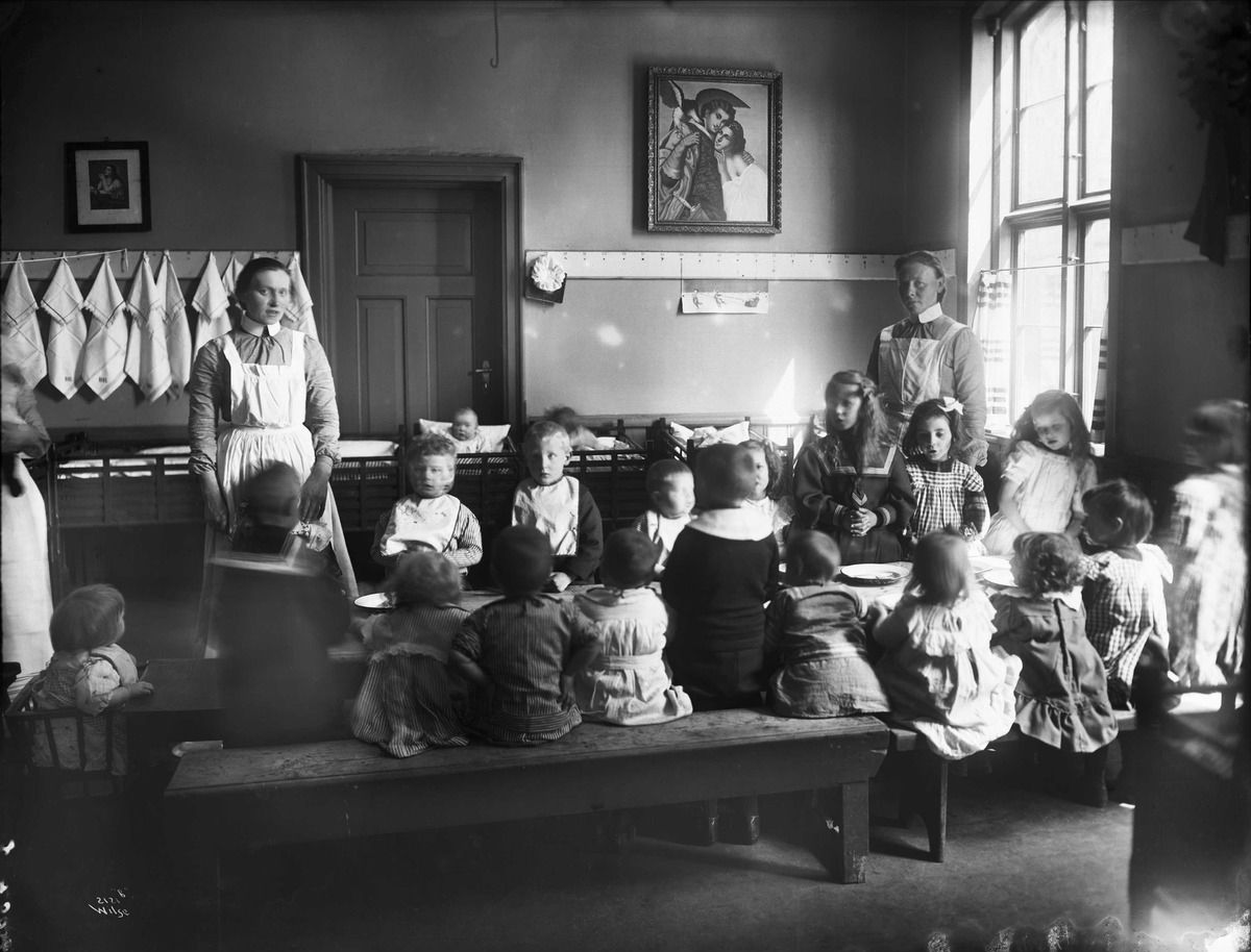 Fotografi av en barnekrybbe drevet av Frelsesarmeen, interiør, med barn og voksne kvinner