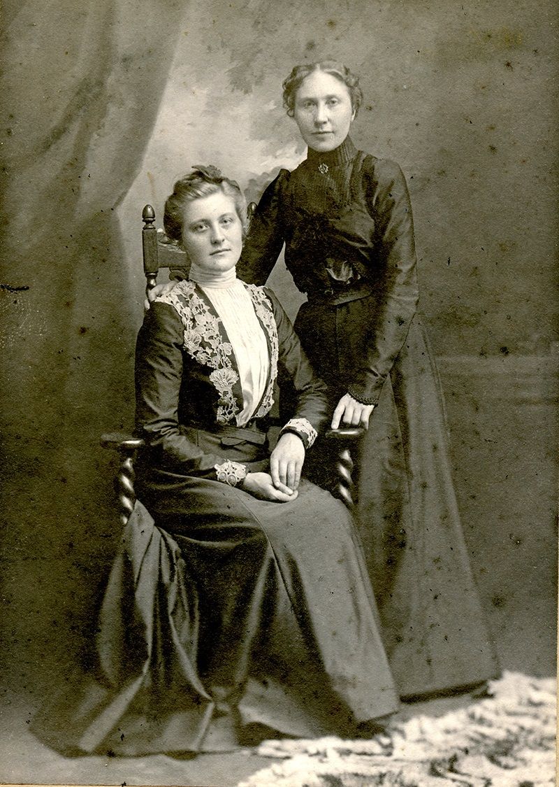 Svart-kvitt foto av to unge kvinner. Begge har lange kjolar. Ei av dei sit og den andtre står ved sida med armen kilande rund skuldra til den sitjande.