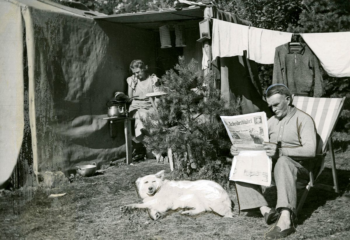 Fotografi som viser campingliv: mann med avisa Arbeiderbladet, kvinne med kaffekjele, hund. Klesvask til tørk.