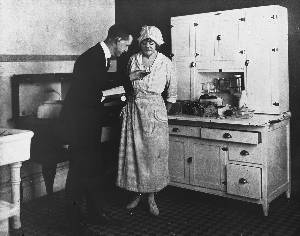 Foto av mann i dress som instruerer husmor, som for øyeblikket står med ryggen til kjøkkenbenk