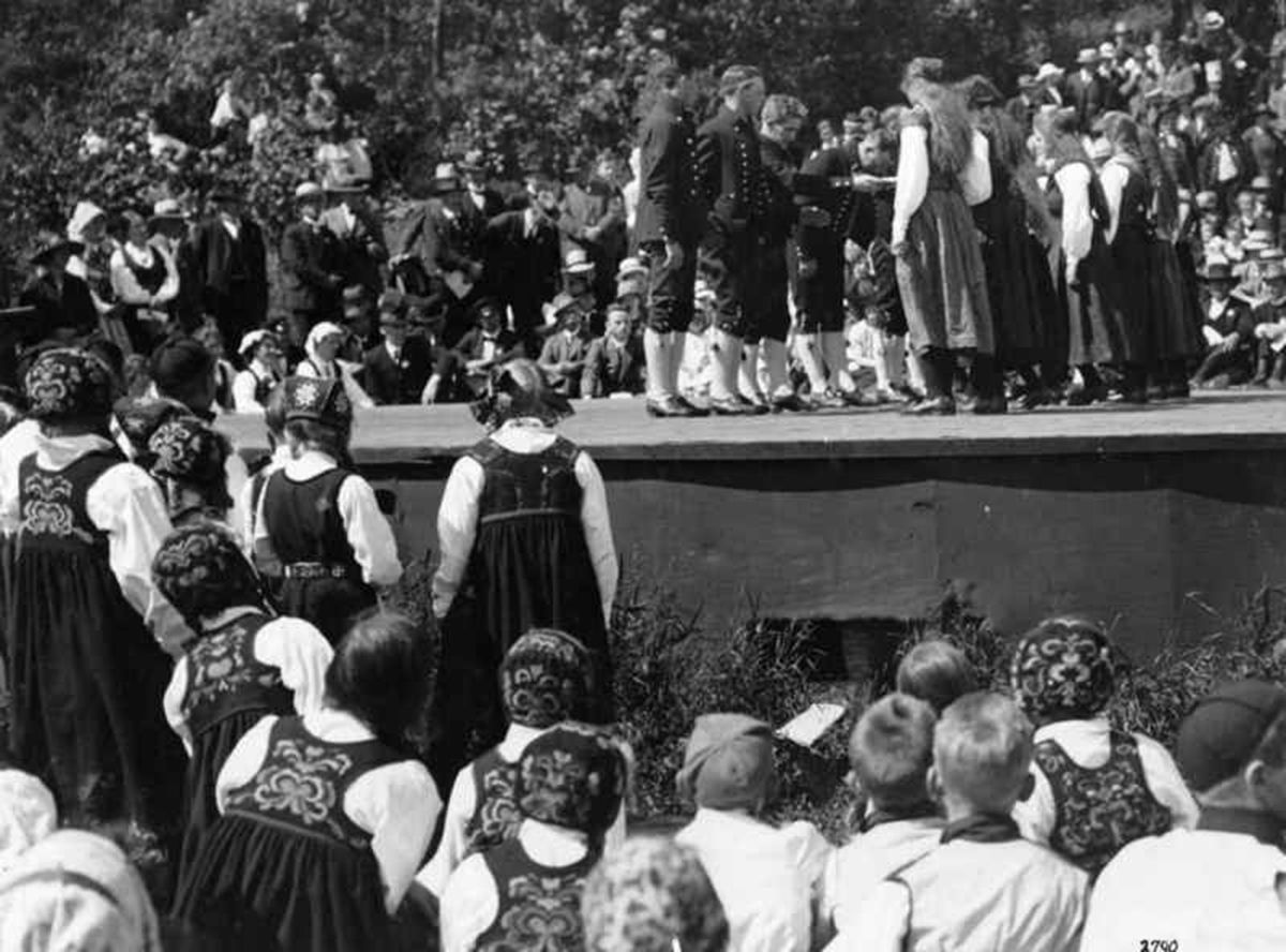 Fotografi som viser folkedans på en scene. Både tilskuere og utøvere ikledd bunad