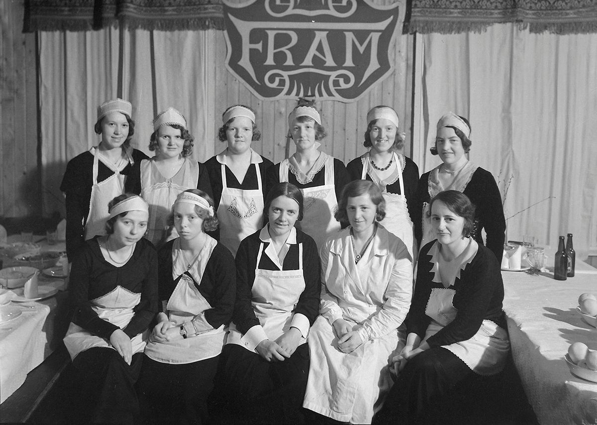 Fotografi som viser kvinner oppstilt i to rekker, med arbeidsklær: forkle, pannebånd, noen med frakk
