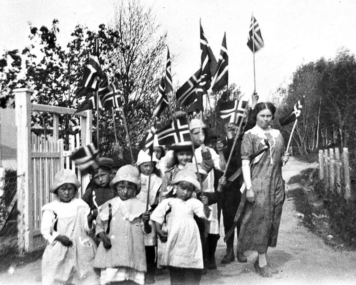 Fotografi av barn og kvinne i 17. mai-tog, gående gjennom en port. Vifter med norske flagg
