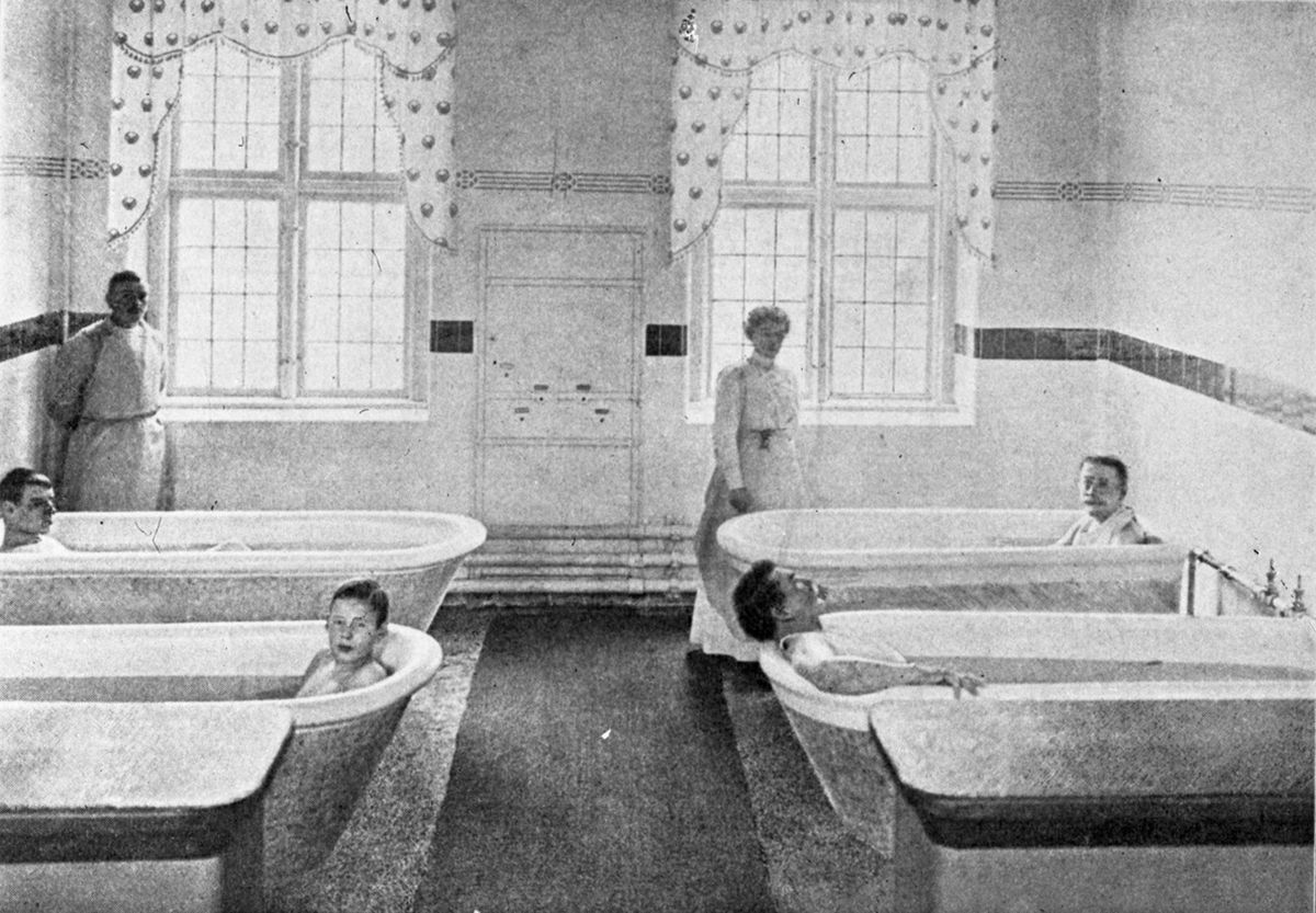 Fotografi som viser rom med fire badekar, mennesker i hvert bad, mann i hjørne og kvinne stående i midten av bildet