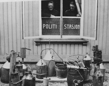 Under brennevinsforbudet ble hjemmebrenning et stort problem i Norge . Her skuer to politibetjenter utover beslaglagte hjemmebrent­apparater utenfor Skedsmo politistasjon. Foto: Akershus arbeiderblad, Akershusbasen