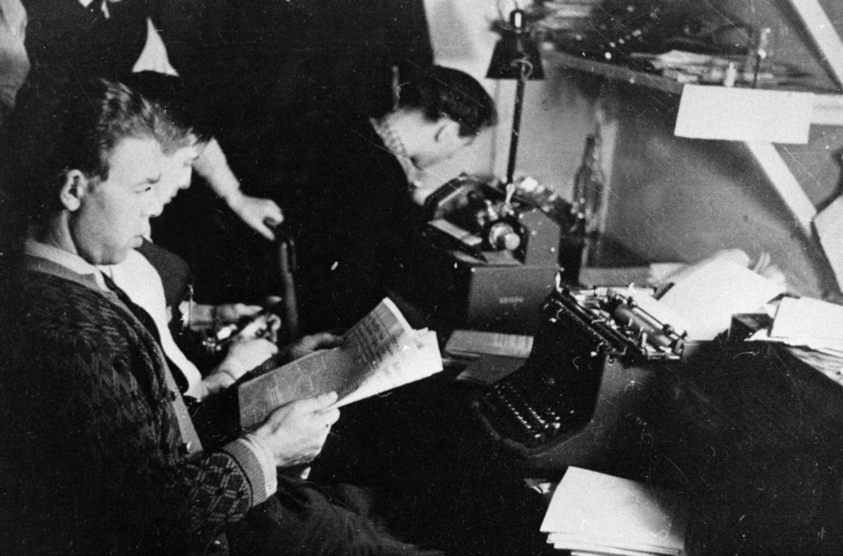 Tre menn sitter trangt foran skrivemaskin og liten trykkvalse
