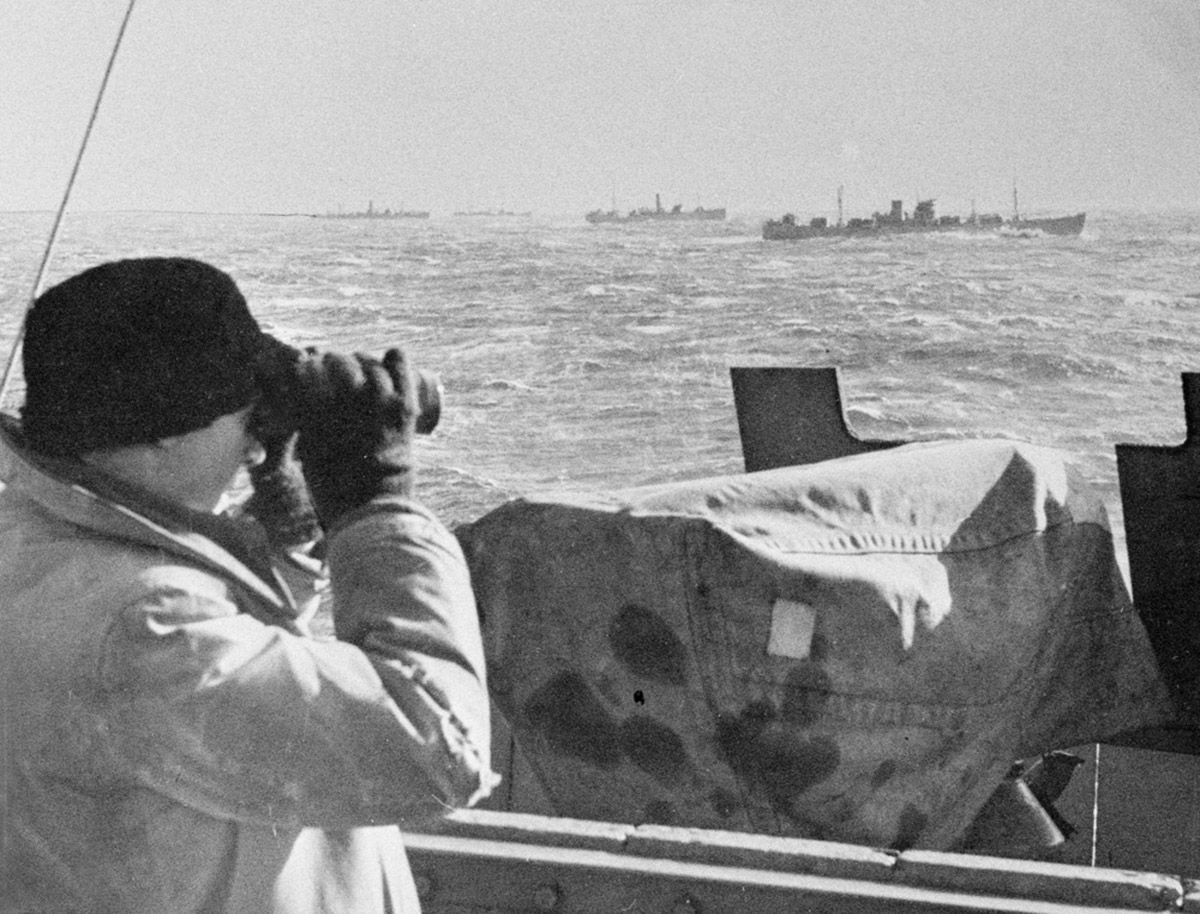 Svart/hvitt foto av mann med kikkert om bord på skip i forgrunn, speider ut mot andre konvoifartøy