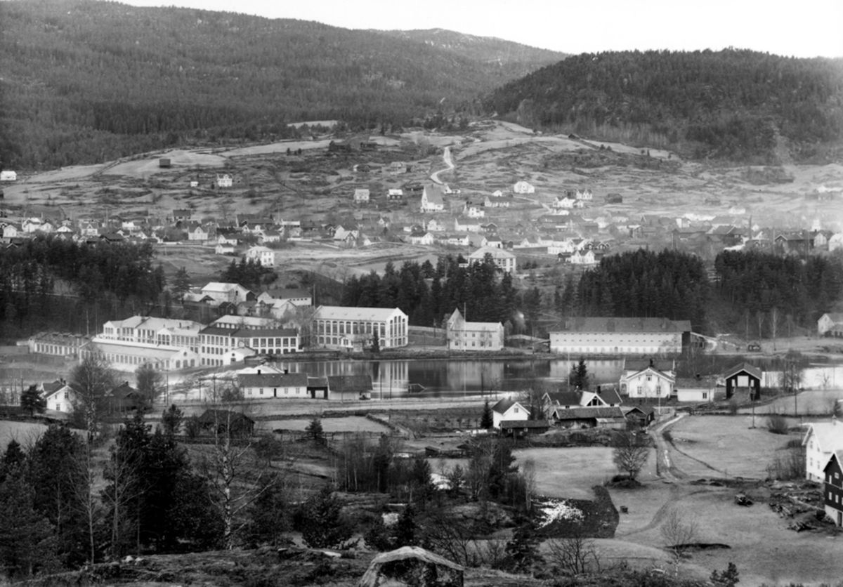 Flyfoto i svart/hvitt av Kongsberg Våpenfabrikk: bygninger ved elv, bygdelandskap i bakgrunn