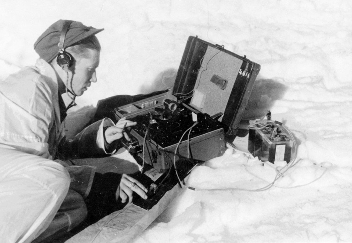 Mann med vinteruniform sitter på huk over en radiosender i snøen
