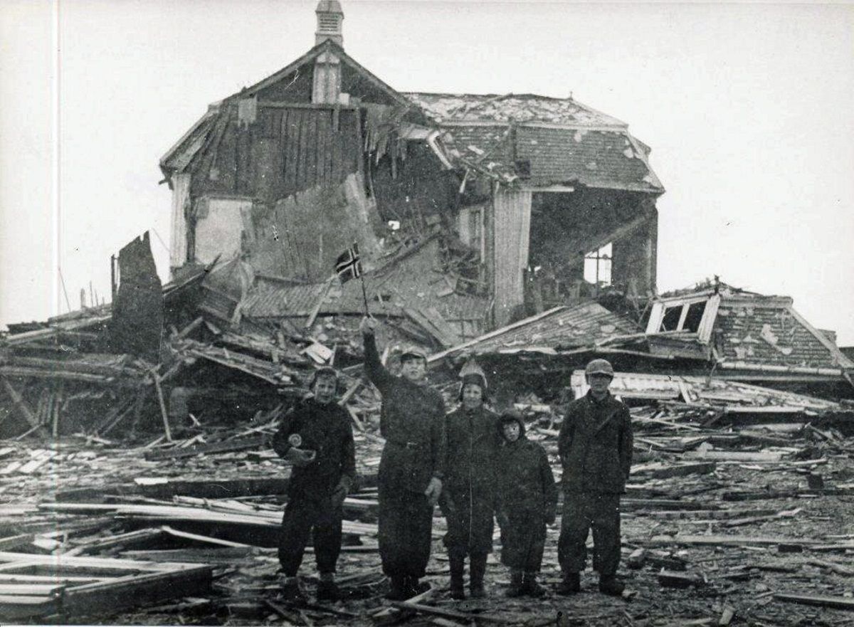 Svart-kvitt-bilete. Fem gutar står føre eit bomba trehus i ruinar. Den eine held eit lite norsk flagg opp i lufta.