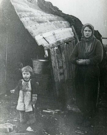 Ikke alle fulgte den tyske evakueringsordren i oktpber 1944. 23 000 ble igjen og en rekke provosoriske hjem ble bygd av materialer som den gjenverende befolkningen kom over. Ødlagte båter ble gjerne til tak og etterhvert dekket man til vekker og tak med jord og torv slik at hjemmet fikk utseende som en gamme. Her er Ingeborg Torbjørnsen og datteren Sigrun foran «båtgammen» som ble hjemmet deres i Mehamn etter nedbrenningen i oktober 1944 . Foto: ukjent/Museene for kystkultur og gjenreisning i Finnmark IKS