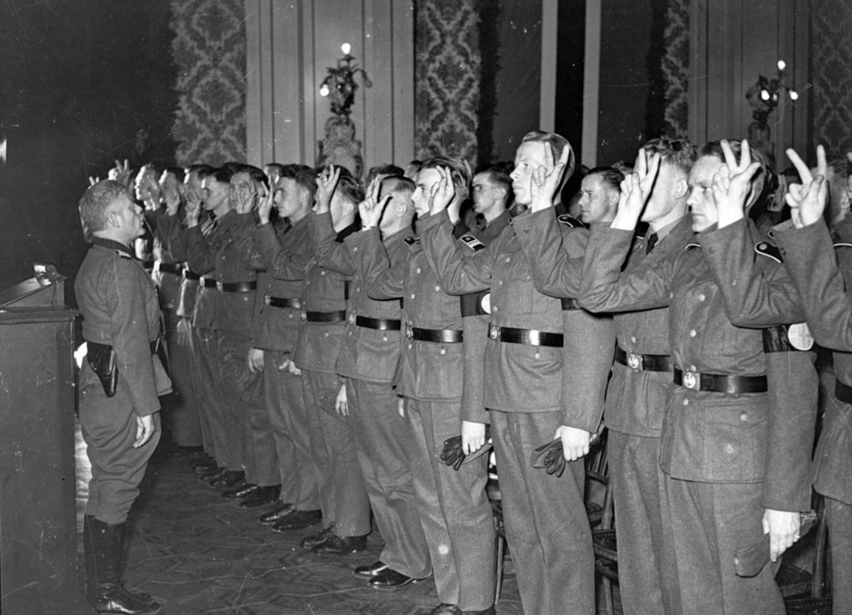 Fotografi som viser menn i uniform på rekke, hevet hånd: edsavleggelse