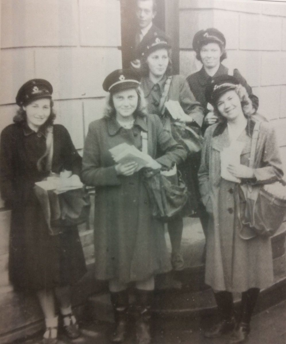 Svart-kvitt-foto av fem kvinner i kåper, postlue og med postveske.