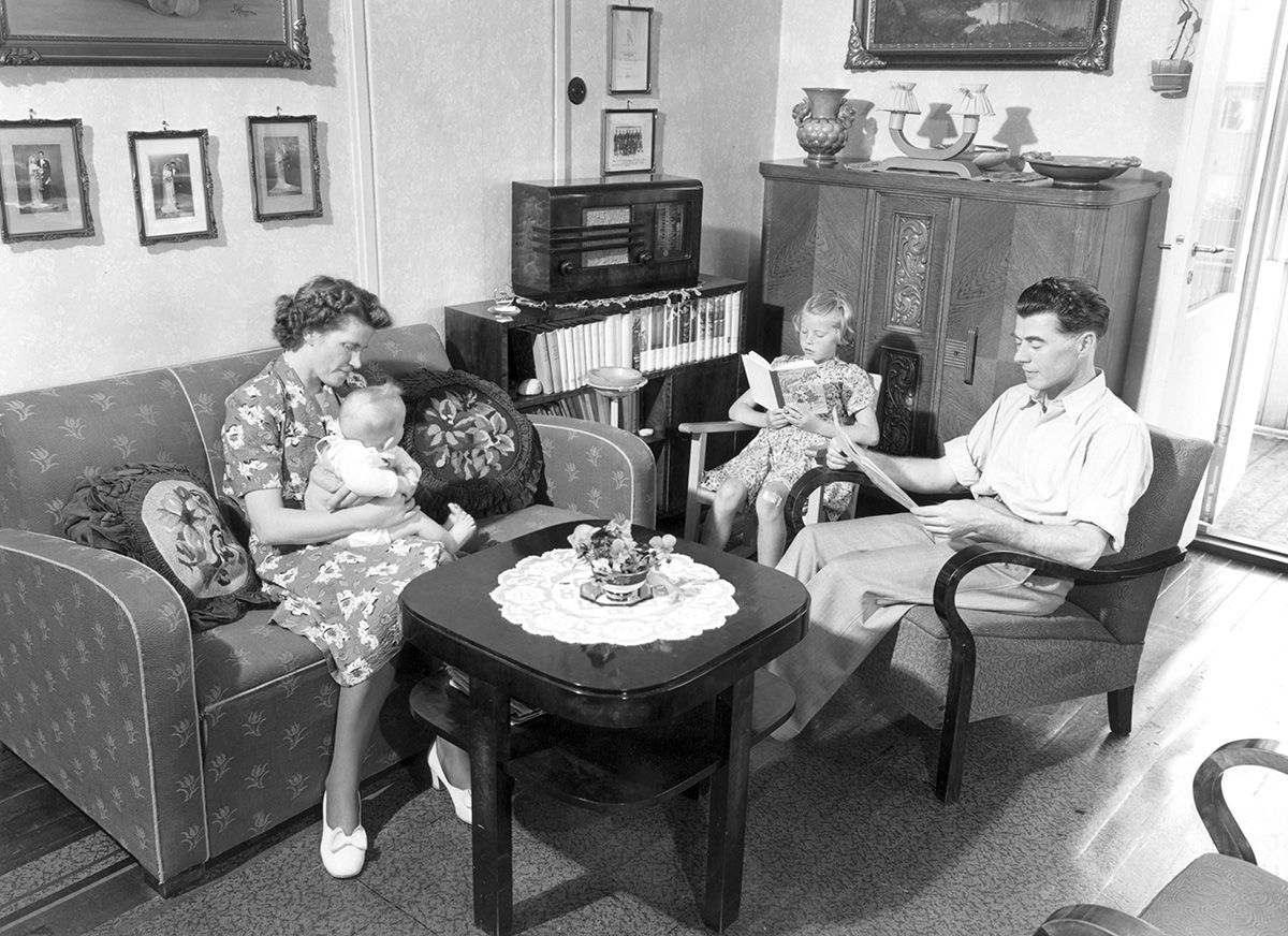 fotografi av familie, interiør: kvinnen med spedbarn på fanget i sofa, mann i stol med avis, jente i stol med bok. 