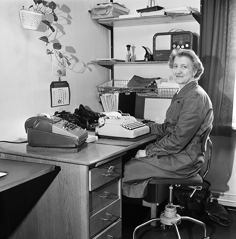 Fotografi, kvinne i arbeidsdress ved kontorpult og skrivemaskin, blikket mot kamera
