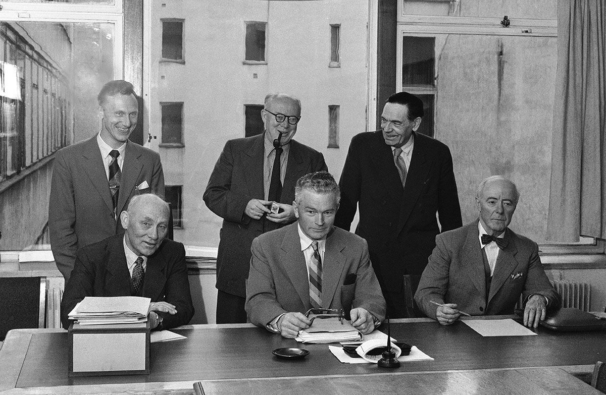 Fotografi av Schei-komiteen: seks menn i to rader, første rad sittende ved skrivebord