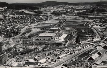 Dette fotografiet fra 1960 viser Standard Telefon- og Kabelfabrikk i midten av bildet, Risløkka til venstre og Alnabanen i forgrunn. Utsyn mot Groruddalen. Kabelfabrikken ble grunnlagt i 1915, og i etterkrigsårene ekspanderte bedriften utover jordene. Les mer om hvordan gaffeltrucken formet bylandskapet . Foto: Widerøes Flyveselskap/Schroder/Oslo byarkiv/ CC BY-SA