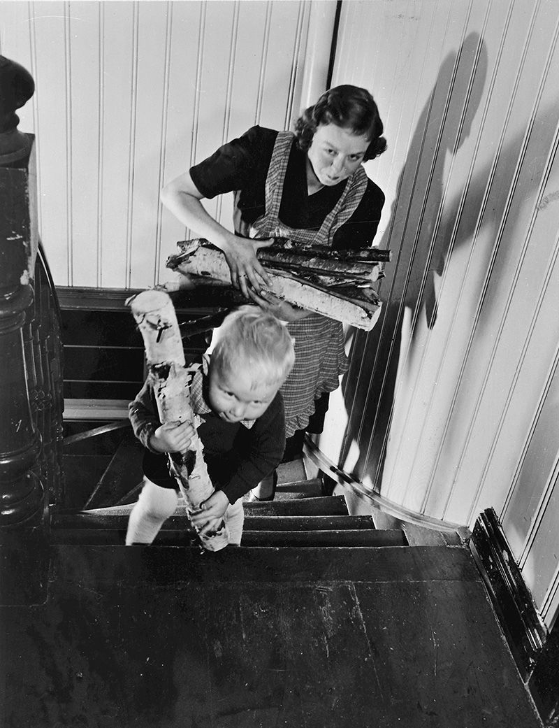 Fotografi fra toppen av en trapp: lite barn med vedkubbe, etterfulgt av mor med vedstabel