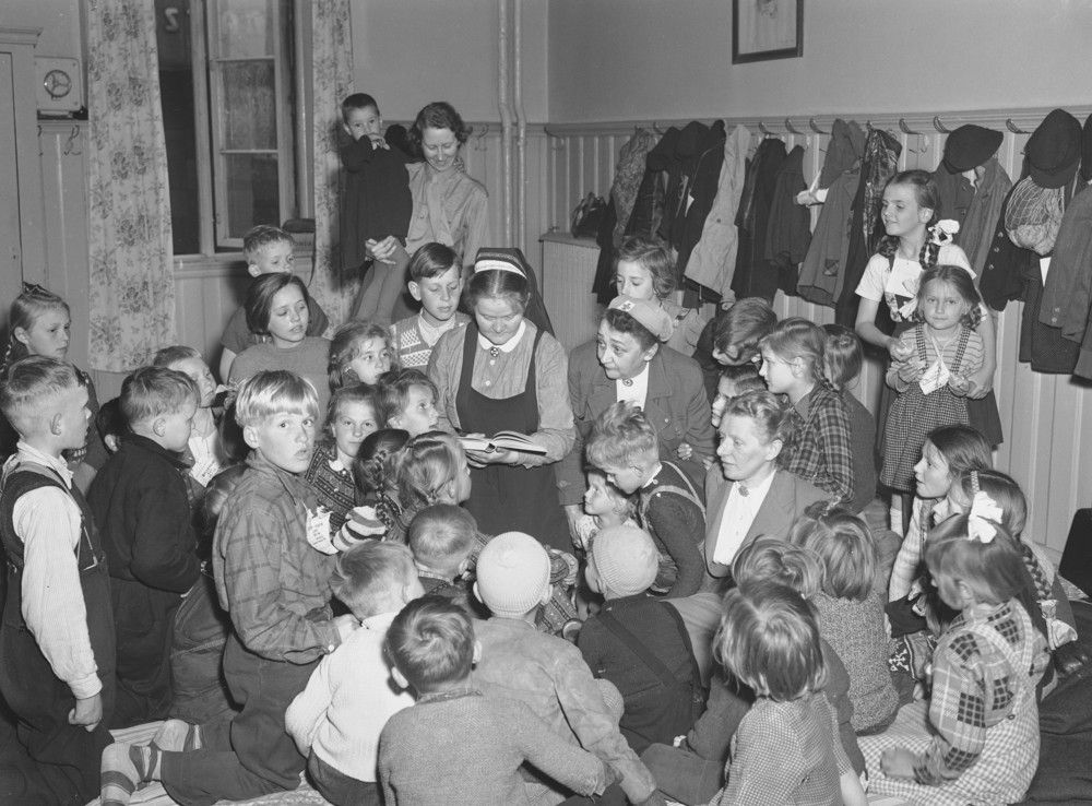 Svart-hvitt foto av flokk med barn rundt kvinne med religiøst hodeplagg som leser