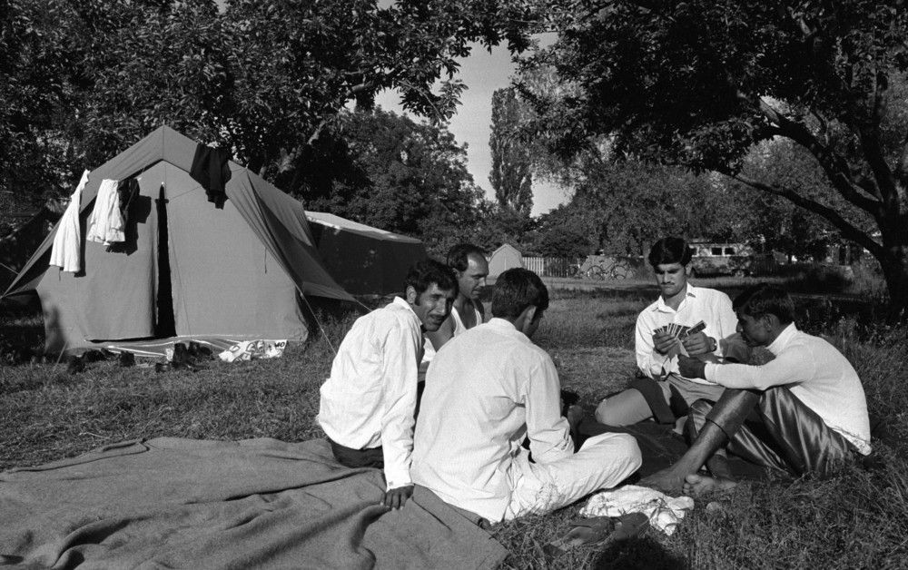 svart-hvitt foto av unge menn som sitter i sirkel på bakken og spiller kort; telt med hengende klær i bakgrunnen