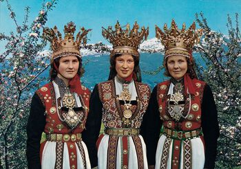 Et kjent motiv som går igjen i norsk tekstilkunst er åttebladsrosen. Denne finner vi ofte som del av bunadsbroderi og hardangersøm, som på vesten til kvinnen i midten på dette postkortet fra 1970-tallet. (Bilde 1 av 3) Foto: Normanns kunstforlag /Nasjonal­biblioteket