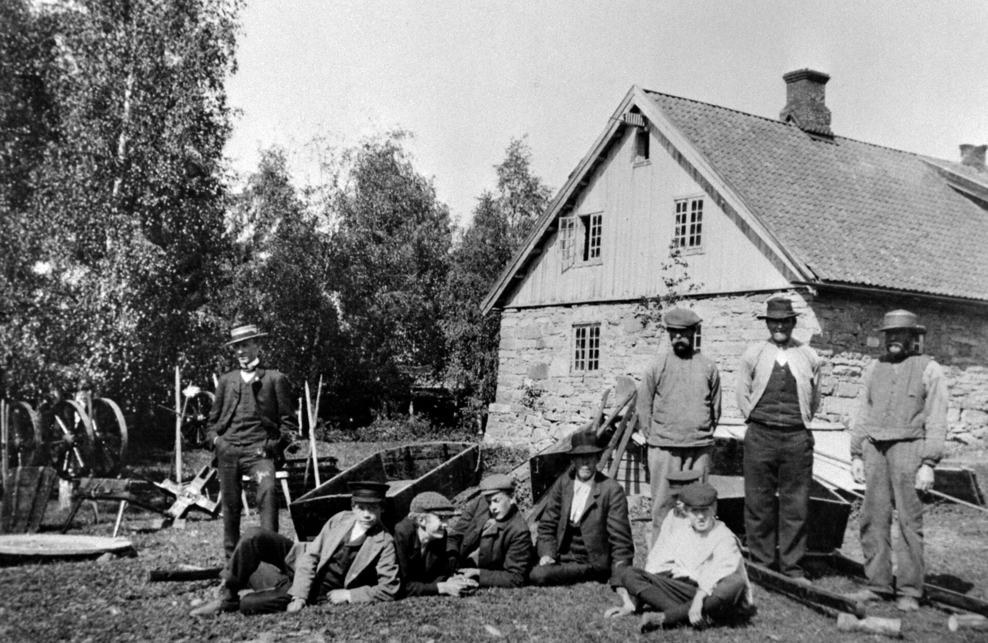 Bildet viser ni husmenn. De eldste står. Hus i bakgrunnen