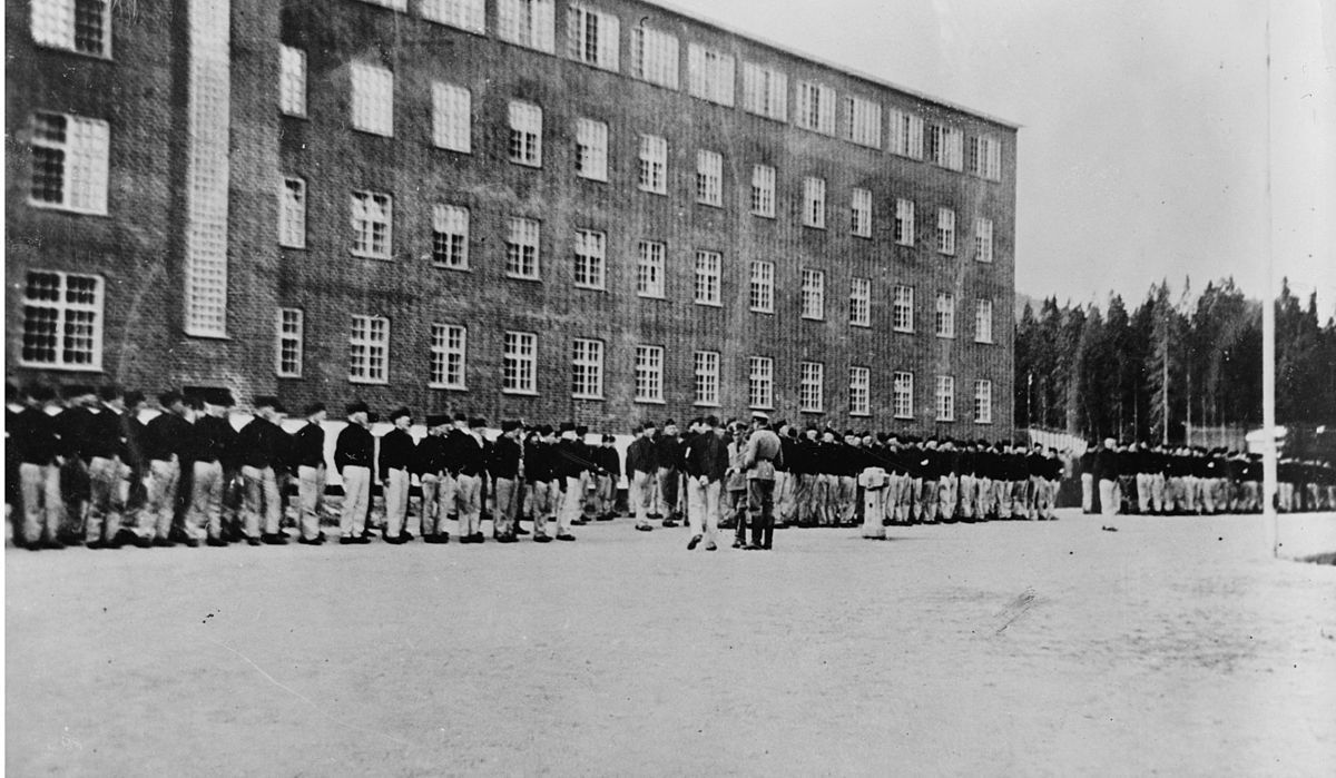 Fotografi av fanger oppstilt utenfor hovedbygningen i Grini fangeleir. 