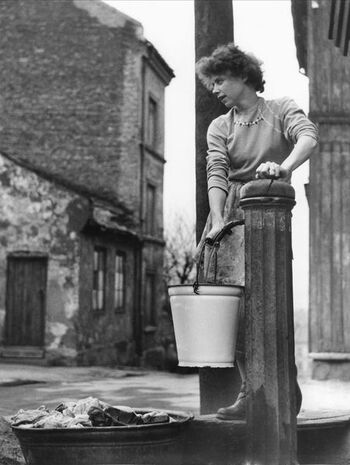 Kvinne ved vannpost på Enerhaugen i Oslo. Fotografiet er fra mellom 1950-55. Ukjent fotograf.&amp;#160; Noen av bygningene som sto på Enerhaugen kan ses i Friluftsmuseet ved Norsk Folkemuseum &amp;#160;på Bygdøy, Oslo.&amp;#160;